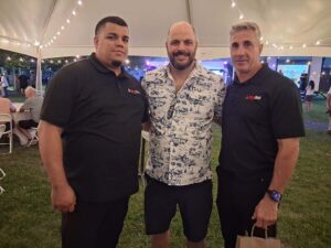 Three men pose together under a large outdoor tent with string lights. Two wear black polos and one wears a patterned shirt and shorts. People are seated at tables in the background.