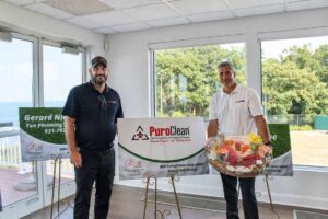 Two men stand indoors by large windows. One holds a gift basket; the other stands beside a PuroClean sign. Sunlight streams in. Two more business signs are displayed behind them. Trees and water are visible outside.