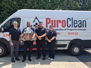 Four people stand in front of a PuroClean van. One person holds a gift basket. All are wearing black uniforms. The van displays the PuroClean logo and contact information. Trees are visible in the background.